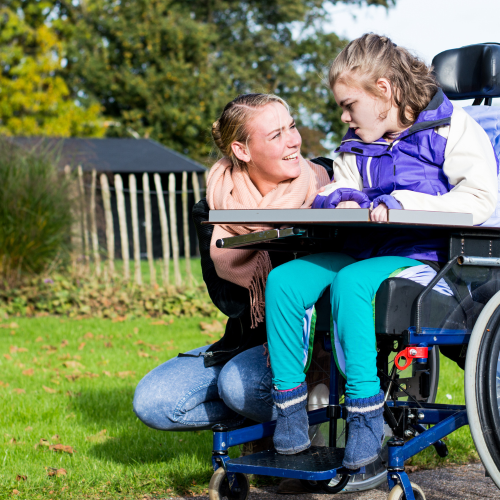 A caregiver speaking with a young wheelchair user outdoors.
