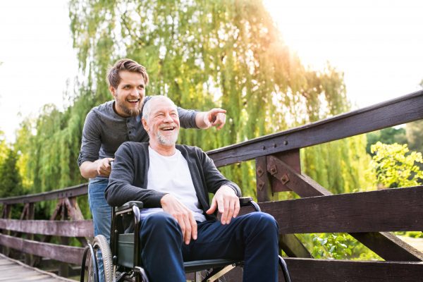 Young hipster son walking with disabled father in wheelchair on wooden bridge at park, pointing finger at something. Carer assisting disabled senior man.